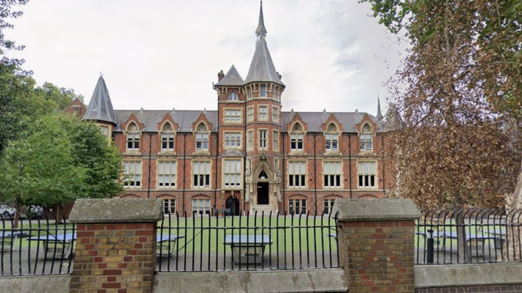 MVPA pictured in its large Victorian red-brick school building with tall arched windows and a central tower, viewed from behind a black metal fence, with trees and an empty courtyard in the foreground.