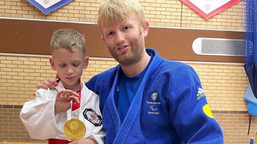 Jacob holding a gold medal standing next to Chris Skelly