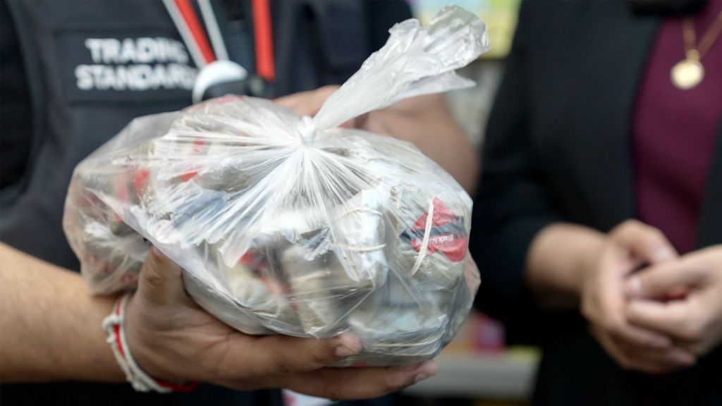 Close-up of a tied, transparent plastic bag filled with empty metal snuff containers being handed between two people. One person wears a dark shirt with partially visible text reading “TRADING STANDARDS,” while another person’s hands and a pendant necklace appear in the background.
