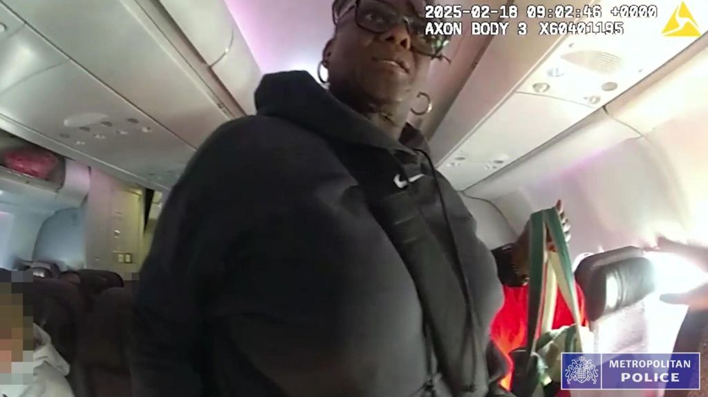 A low-angle body-cam shot inside an airplane aisle, showing a Janice Nix in a black hooded sweatshirt and glasses holding a striped tote bag. A Metropolitan Police logo is in the corner.