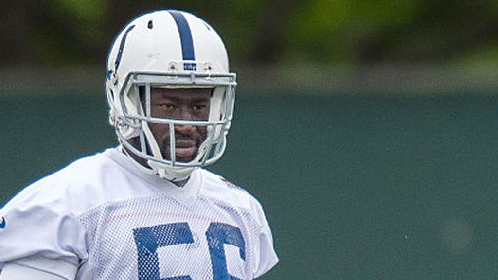 He is wearing his American football uniform, but the image is cut just below his shoulders. He is wearing a white helmet with a blue stripe down the middle and a white tshirt with blue detailing