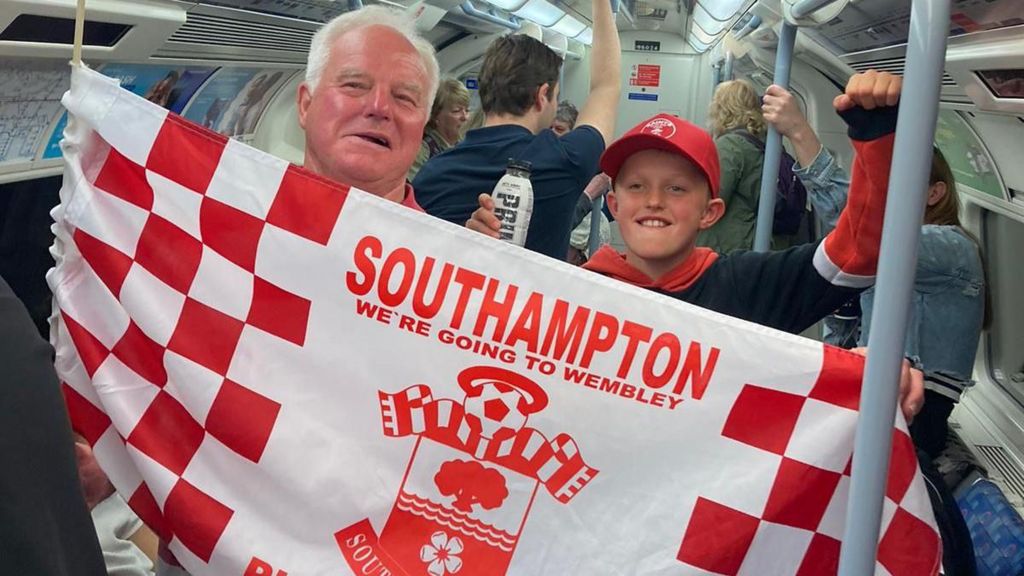 older man and younger boy hold Saints flag on tube