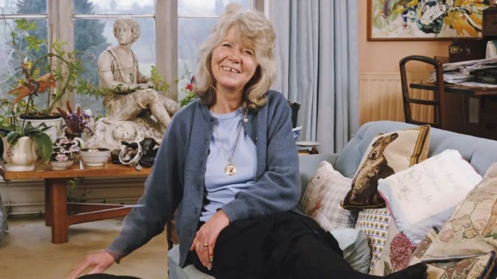 Jilly Cooper sat on her couch smiling at the camera while wearing a dark blue sweater