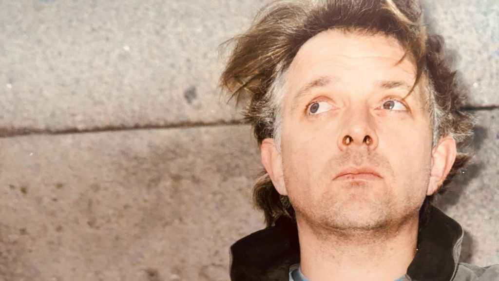 Comedian and actor Rik Mayall, standing with his back against a concrete wall. His hair, mid-brown with grey patches on the sides, is unkempt and he has a five o'clock shadow. He is looking up and behind the camera.
