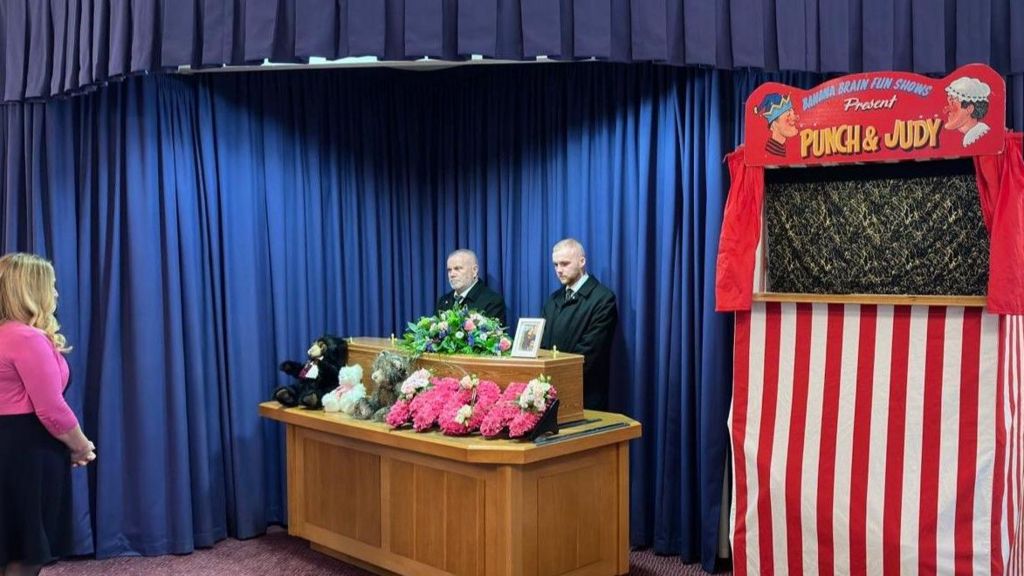 A woman faces the coffin, which has flowers on it and in front of it, as well as teddy bears. A Punch and Judy tent is to the right of the coffin.