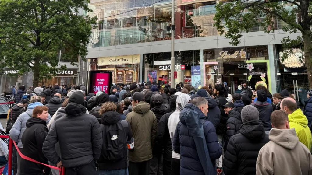 Hundreds of people wait in a queue snaking back from the shop entrance on a pedestrianised street. Glass fronted stores line the road on both sides.