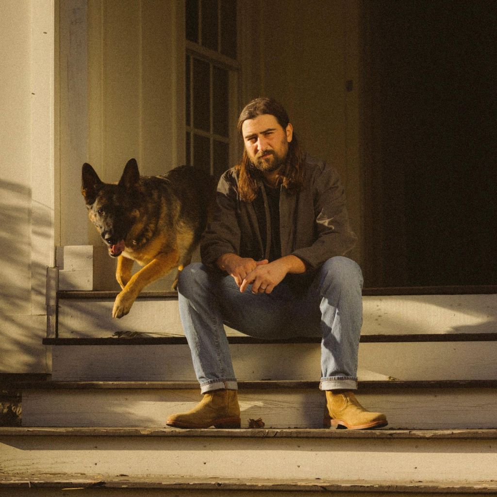 Noah Kahan poses on the porch of a rural house in America, as a dog bounds down the steps
