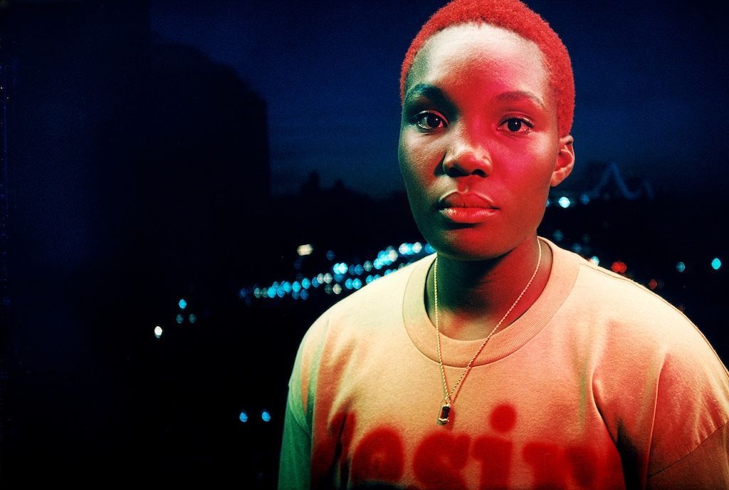 Arlo Parks is athed in red light, as she poses against the soft-focus lights of a city at night-time