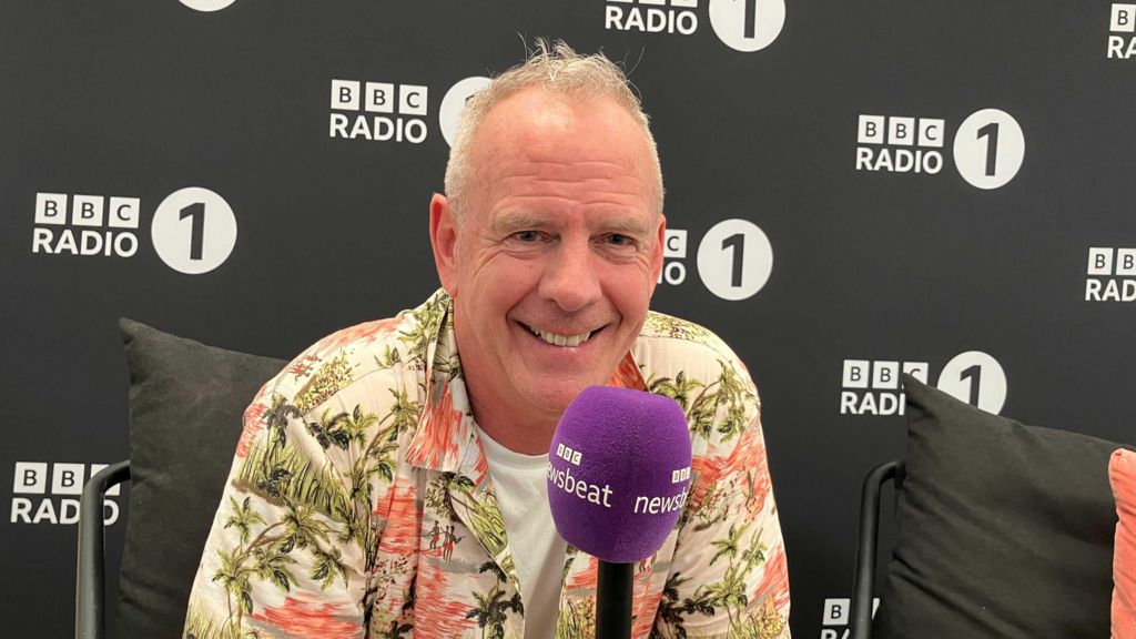 Fatboy Slim smiles holding a purpose Newsbeat microphone. He is wearing an unbuttoned Hawaii shirt over a white t-shirt. He is in front a black backdrop, with the Radio 1 logo on. 