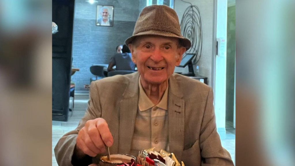 An elderly man smiles (right) for a photo in a cafe. He is wearing a pork-pie hat and a cream jacket and light shirt. He is stirring a hot drink in a mug on a table in front of him which contains a mental container of condiments