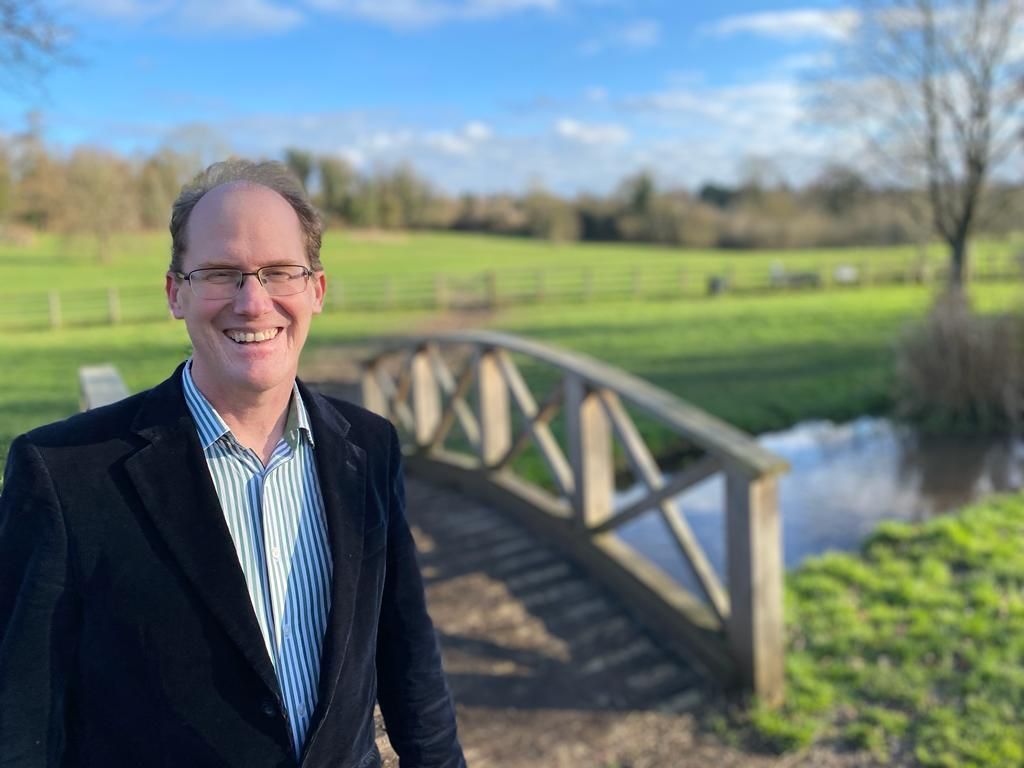 Labour candidate Tim Starkey in a dark suit and shirt, with a bridge and field in the background