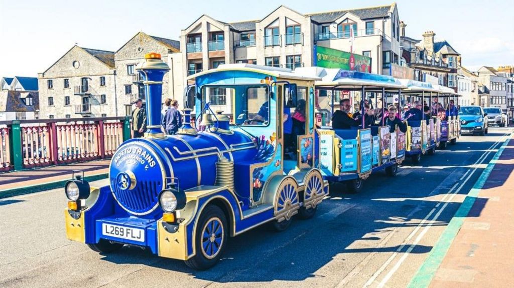 An image of a colourful land train with visitors on board going across a bridge in Weymouth