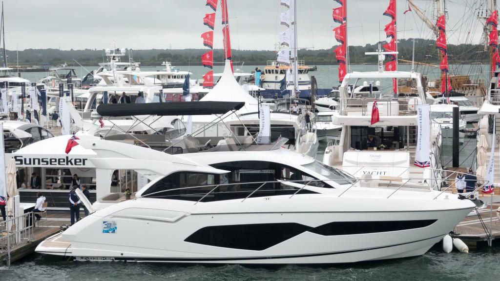 A large white yacht moored up to a pontoon in a busy harbour, next to a marquee with the Sunseeker logo on it
