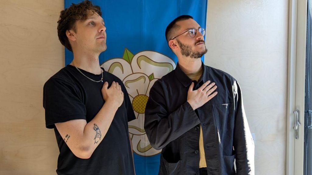 Two young men stand in front of a Yorkshire flag - a white rose on a blue background - hung on a wall. Each has his hand placed across his chest, looking off-camera at the sky. It's a tongue-in-cheek pose reminscent of football players lining up for their national anthem before a game.