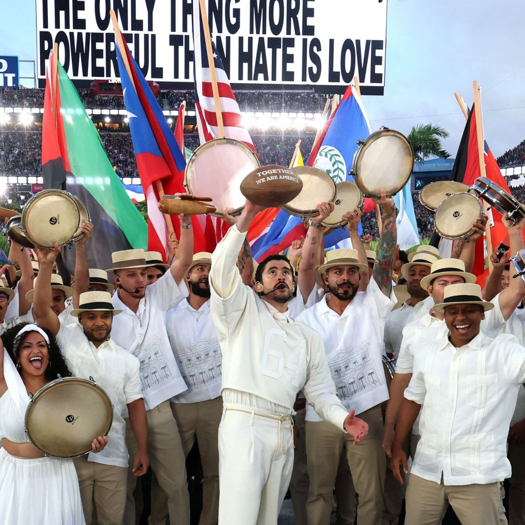 Bad Bunny holding up a football with white writing, surrounded by musicians and dancers holding up instruments and flags
