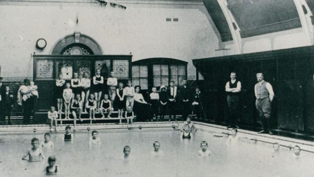 An early black and white picture of the inside of the baths. A number of children in swimming costumes are pictures sitting on benches next to the pool, with many also in the water. 