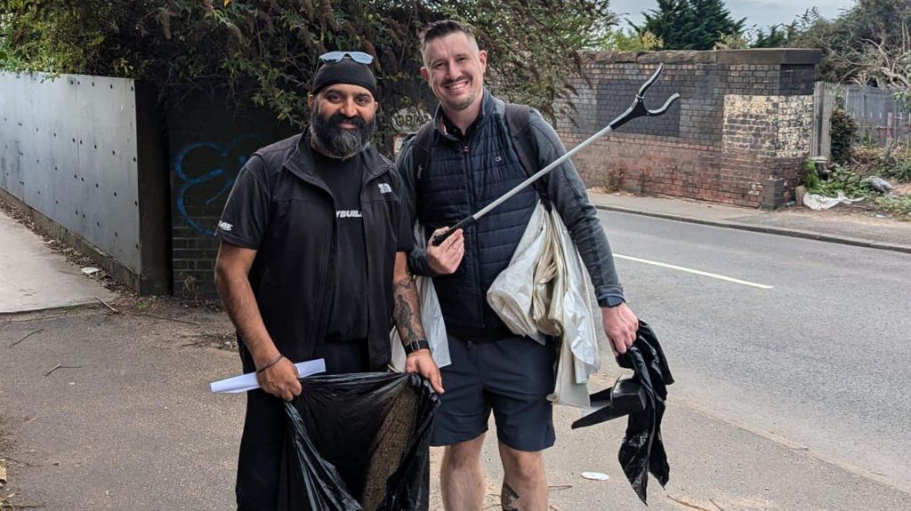 Sukh Singh wearing a black top standing next to Paul Delves who is wearing a black coat. They are standing on the pavement holding rubbish bags and litter pickers.