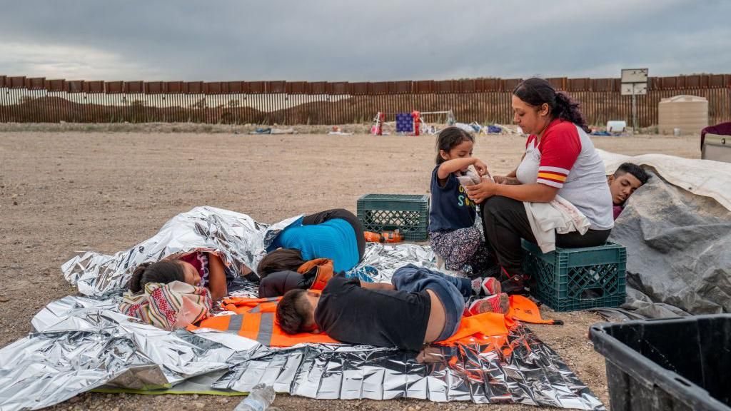 Children sleeping on ground, mother sitting on milk crate while talking to daughter