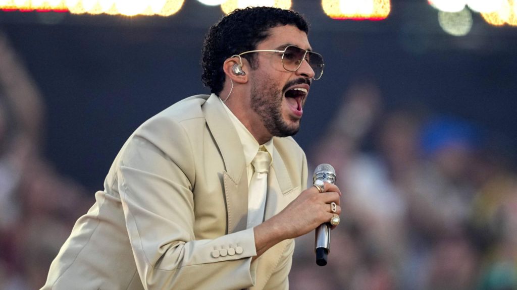 Bad Bunny in dark glasses, a beige suit, and cream shirt and tie singing into a microphone on stage during a concert in Barcelona on 22 May 2026