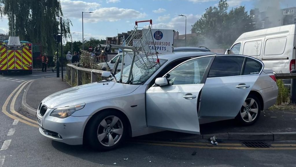 A silver BMW on the pavement at a road junction, with a shopping trolley mounted on the windscreen. The doors are open and wisps of smoke can be seen in the background