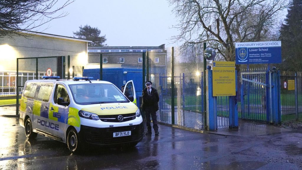 A police van is parked inside the gates of Kingsbury High School in Brent at dusk, while an officer stands nearby on a wet road with school buildings and signage visible.