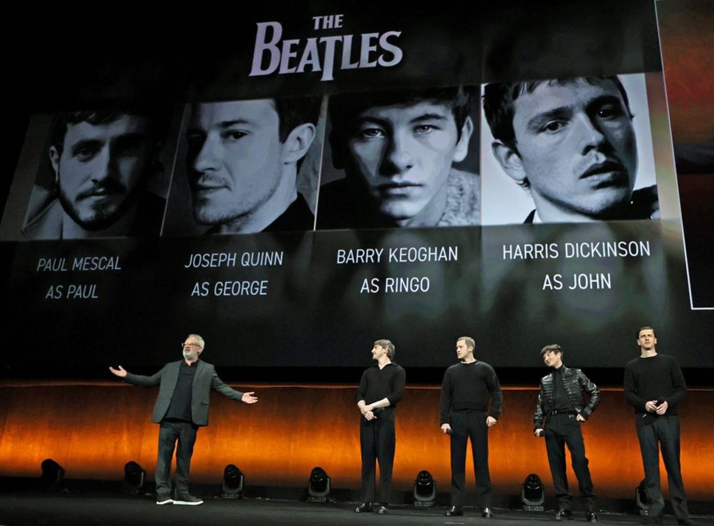 Sir Sam Mendes on stage with the four actors in front of a giant screen with The Beatles logo and black-and-white pictures of the actors above captions stating their names and roles