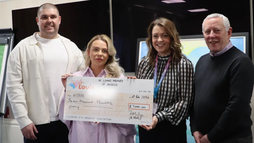 Michael and Alice Miles, and two other people, are holding up a large orange cheque for £7,000 for 4Louis. They are smiling at the camera. Michael Miles, on the very left, has cropped dark hair and is wearing a white jacket and white shirt. Alice Miles, on his right, has long blonde hair and is wearing a pink shirt. Words at the top of the cheque read: 
