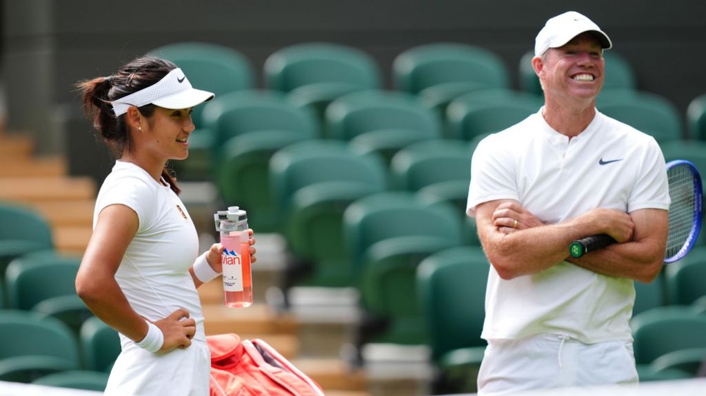 Emma Raducanu, a young female tennis player, with her coach Mark Petchey, talking in a break from a game. She is holding a container full of a pink liquid.