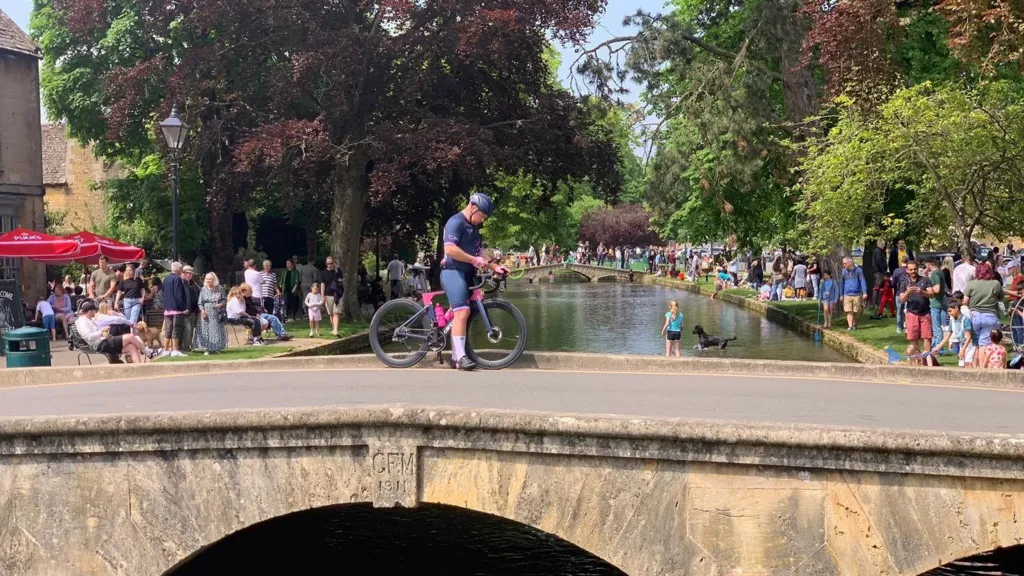 A small Cotswolds-stone bridge with a cyclist wearing Lycra stopped in the middle. The bridge goes over a picturesque river with crowds of people on either side sitting on the grass.
