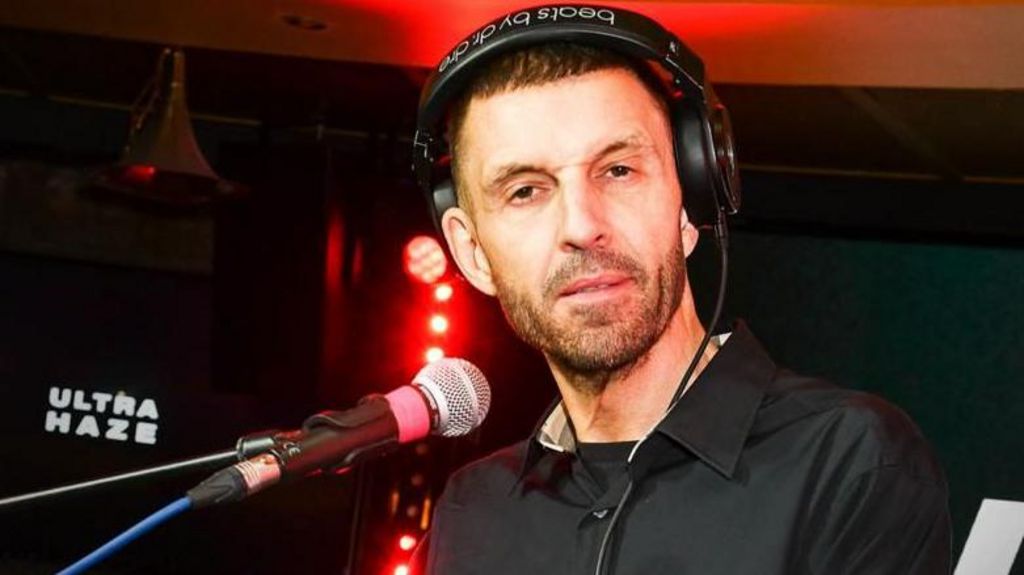 Tim Westwood, with a black shirt on, wearing headphones with a microphone in front of him - there are red studio lights behind him and a black backdrop with the words "ultra haze" 