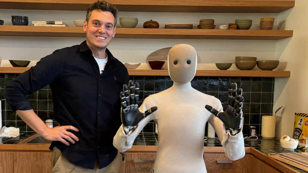 NEO the humanoid robot from 1X and reporter Joe Tidy pose for a photo. Neo is a 5ft robot with soft coverings over it's robot body.