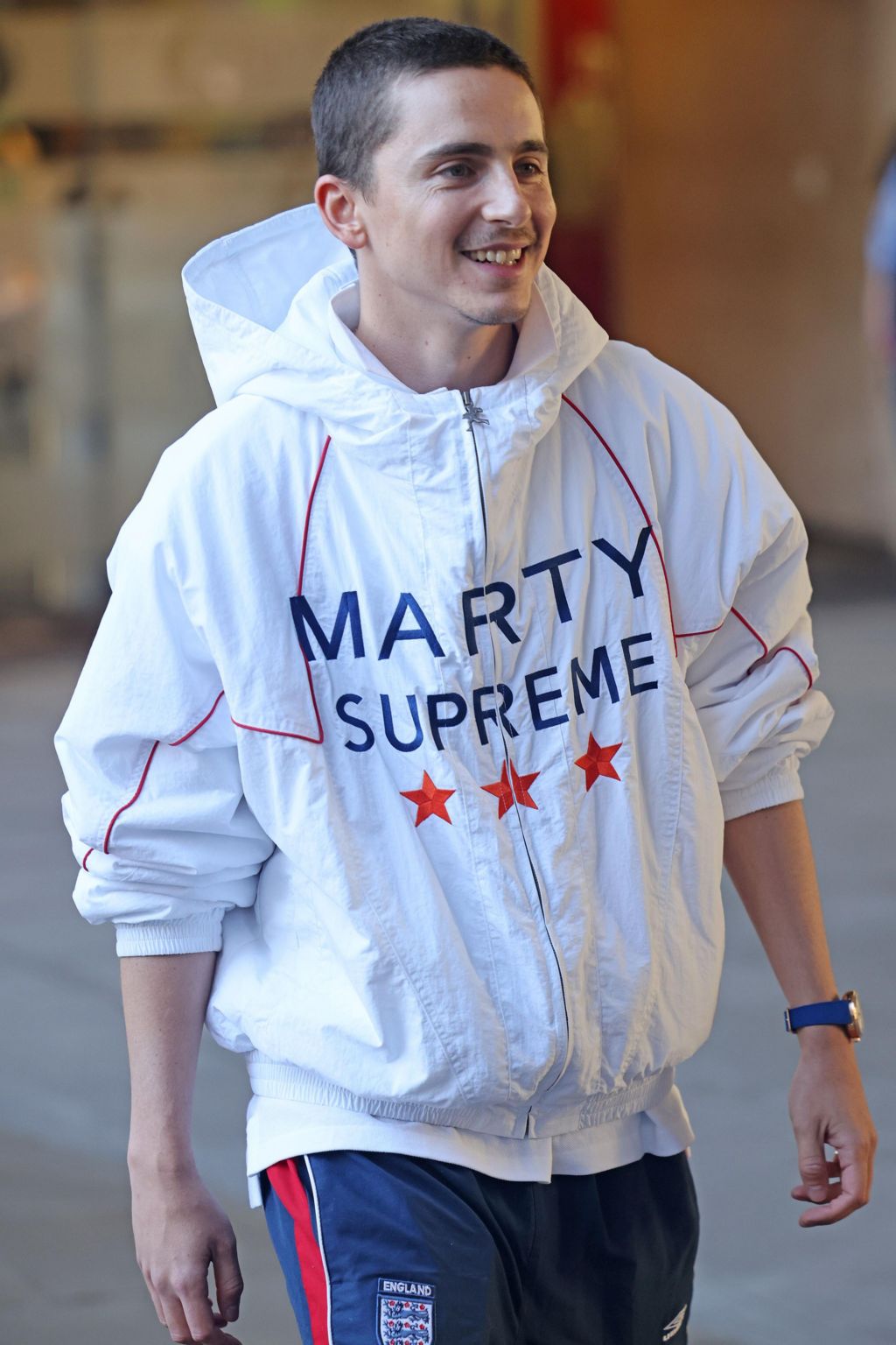 Timothee Chalamet in a white jacket with the name Marty Supreme in large letters and three orange stars on the front