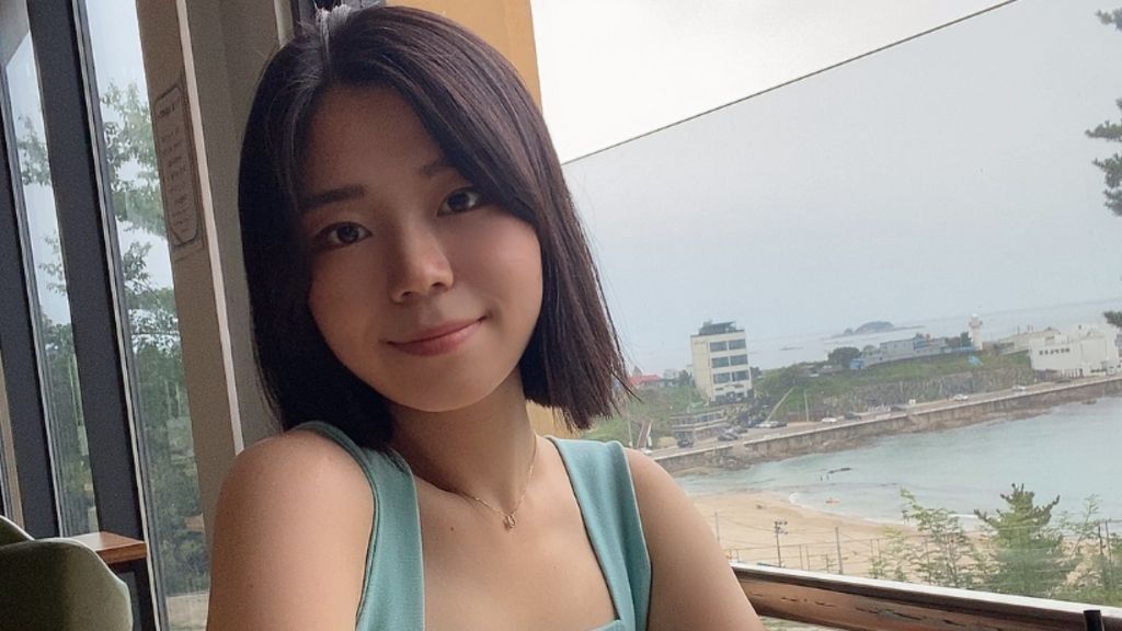 Kim Mi-rim, who has a short dark brown bob and wears a blue vest top, sits in front of a window which looks out onto a seafront