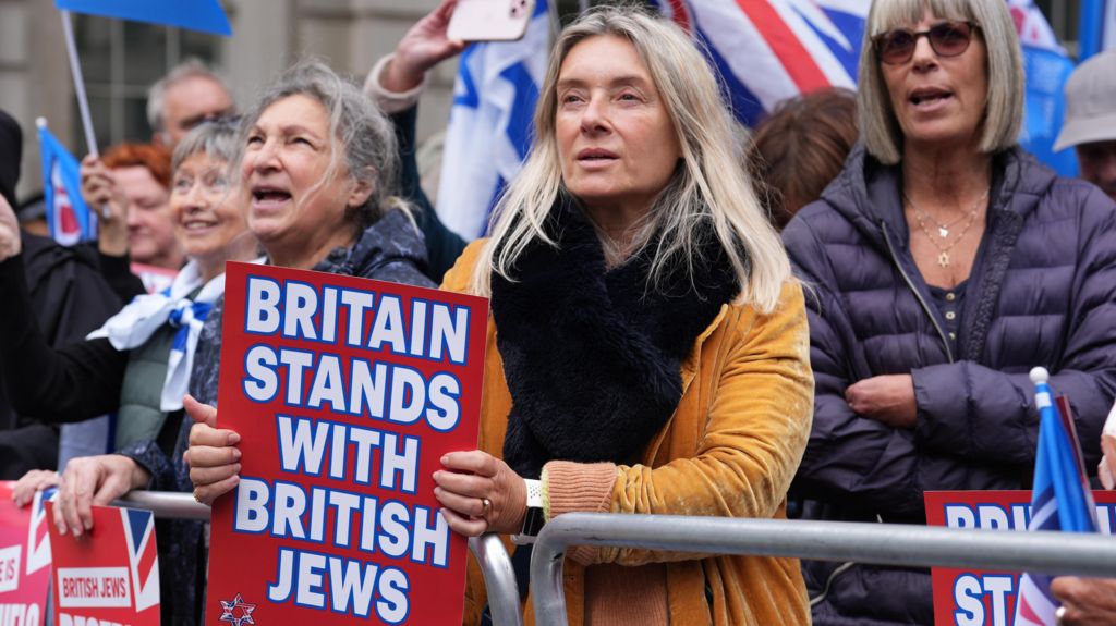 A woman stands at a rally wearing a mustard-coloured velvet coat and black scarf - she holds a sign saying 'Britain stands with British Jews'. A crowd of people can be seen behind her, some with Union flags and Israel flags 