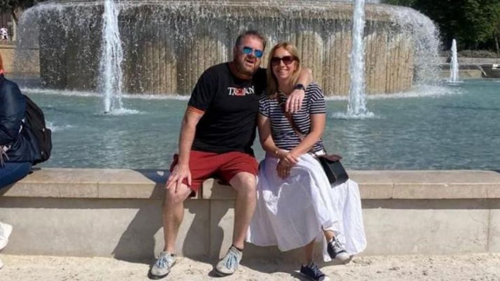 A man and a woman sit on a wall in front of a fountain. 