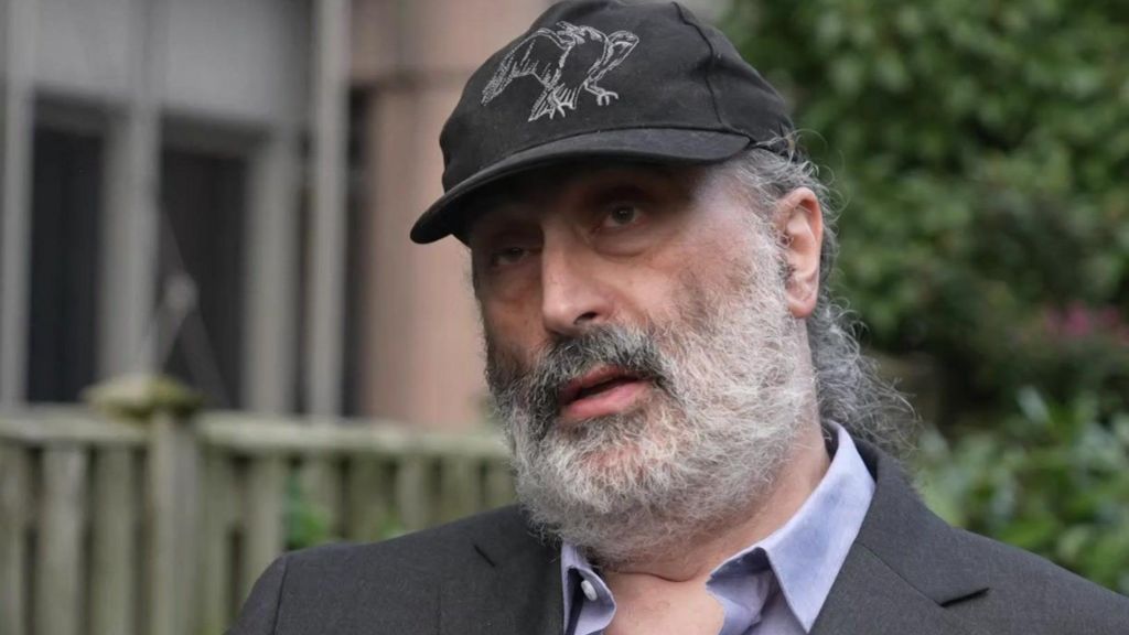 Mark Nygate, a man with a grey beard, wearing a dark suit and a baseball cap.