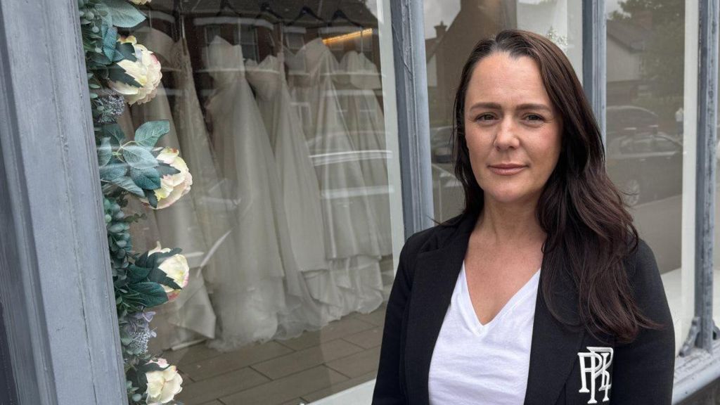 Lauren Elizabeth standing in front of the window of her bridal shop with dresses on display on a rail behind her 