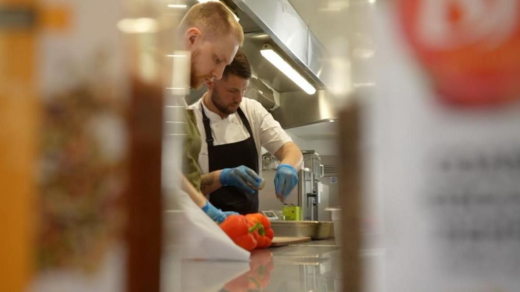 Two chefs in white overalls and aprons working in a stainless steel kitchen 