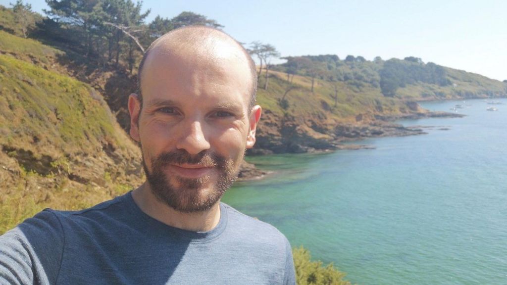 Sam Burns wears a blue T-shirt and has balding, dark hair and a dark goatee beard. He stands in front of a lake with hills to the side of it with trees dotting them