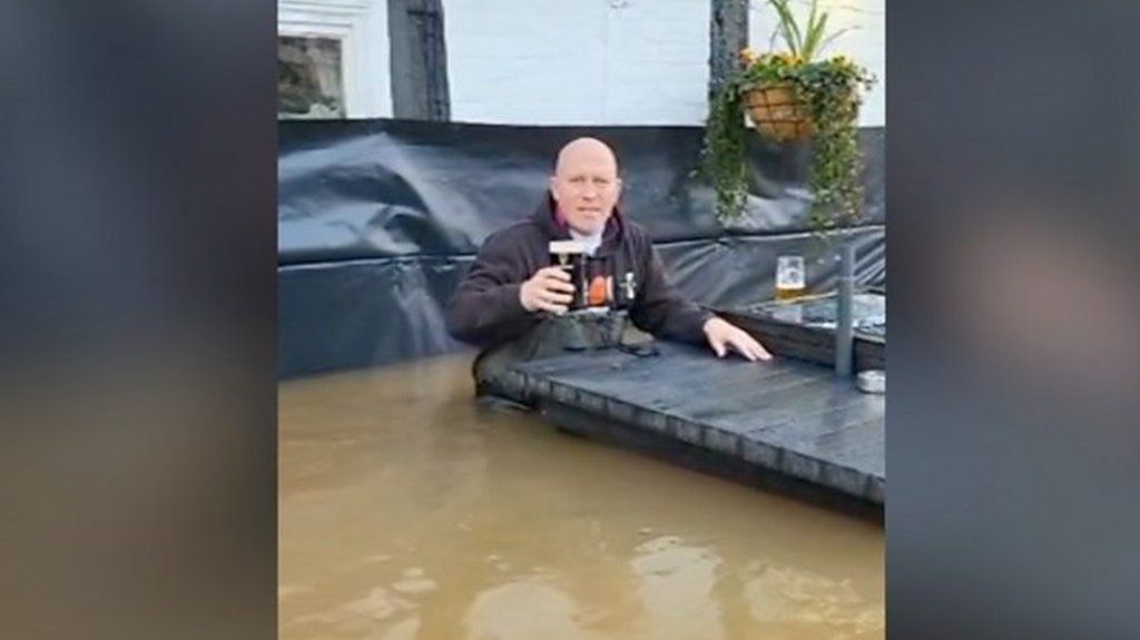 A man sitting in water that is up to his hips holding a pint of beer