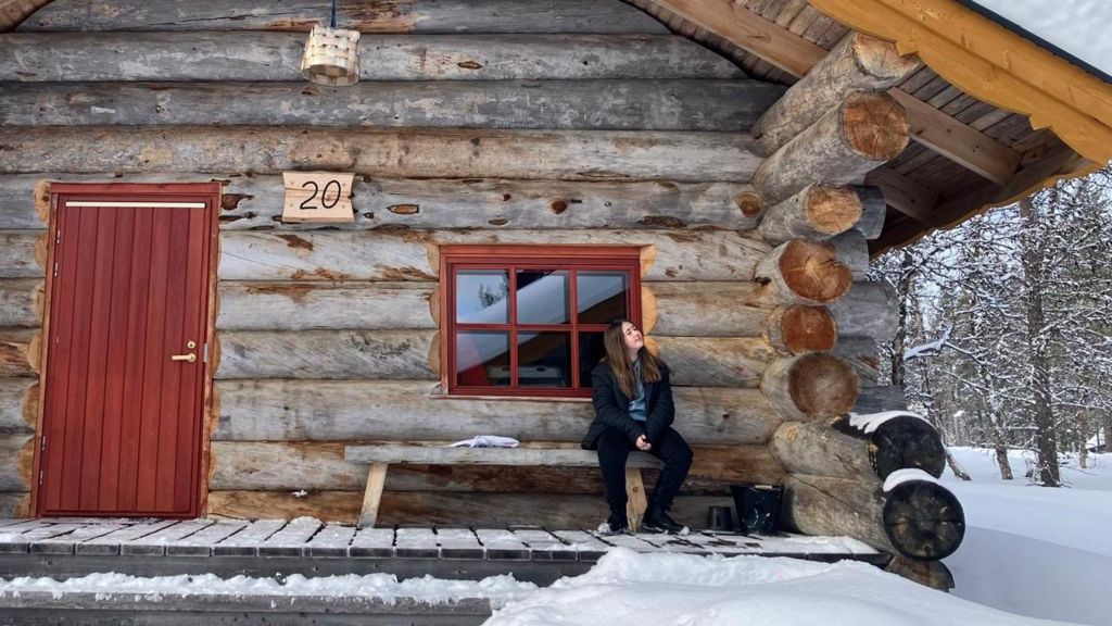 Young girl sits outside a log cabin on a wooden bench, surrounded by deep snow