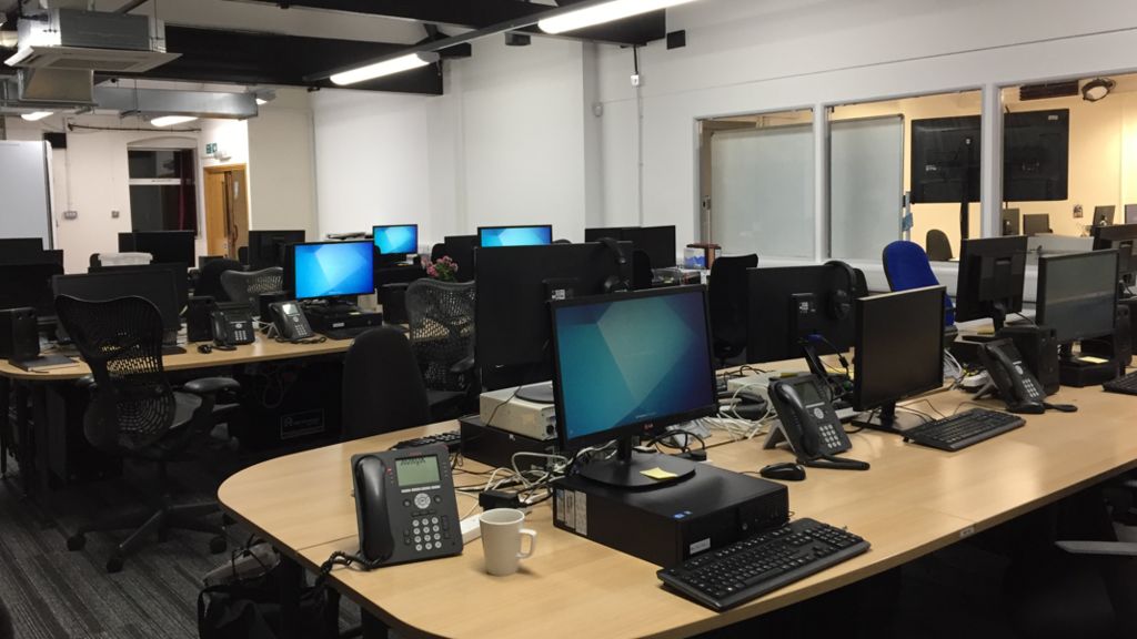 Two rows of light wood desks in an office, with a number of computers and telephones on each desk.