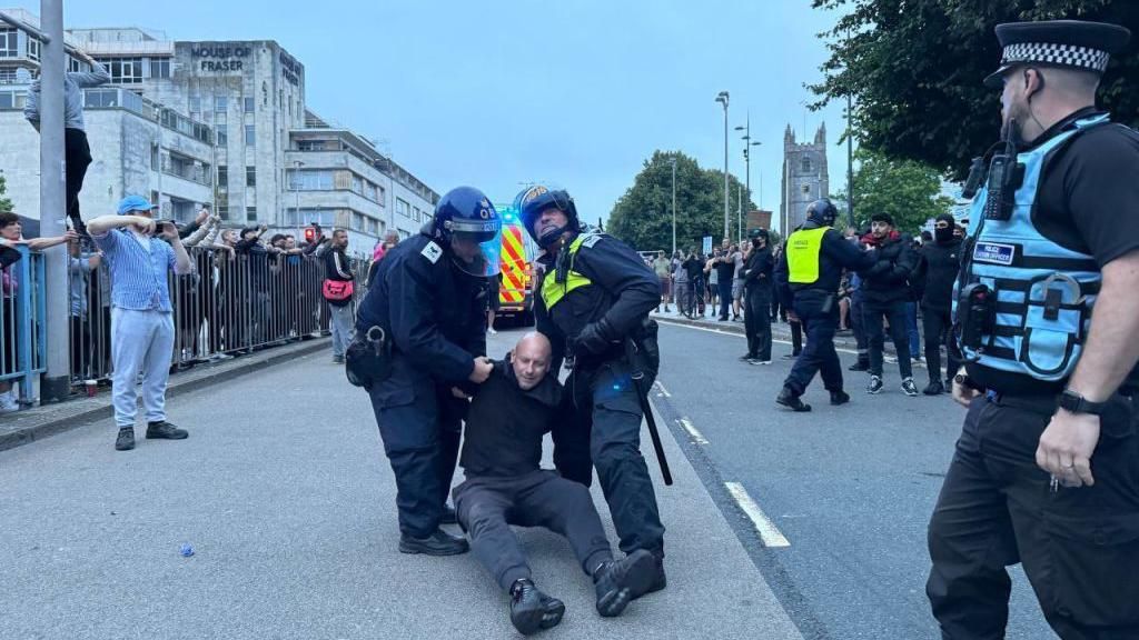 Two police officers in riot gear hold a man sitting on the ground with a police van in the background as protesters stand either side of Royal Parade in Plymouth