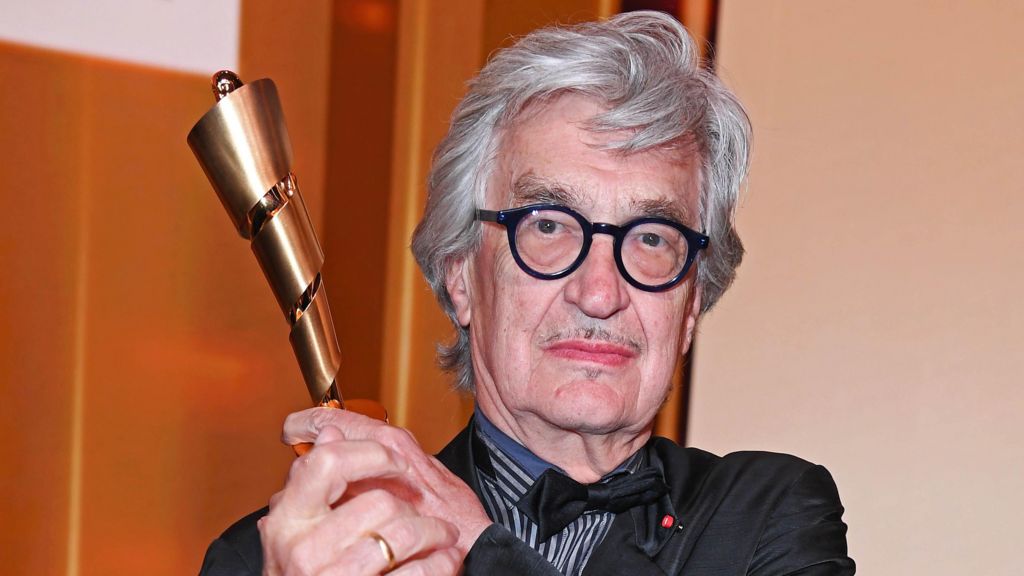 Wim Wenders with grey hair and black glasses holding up an award