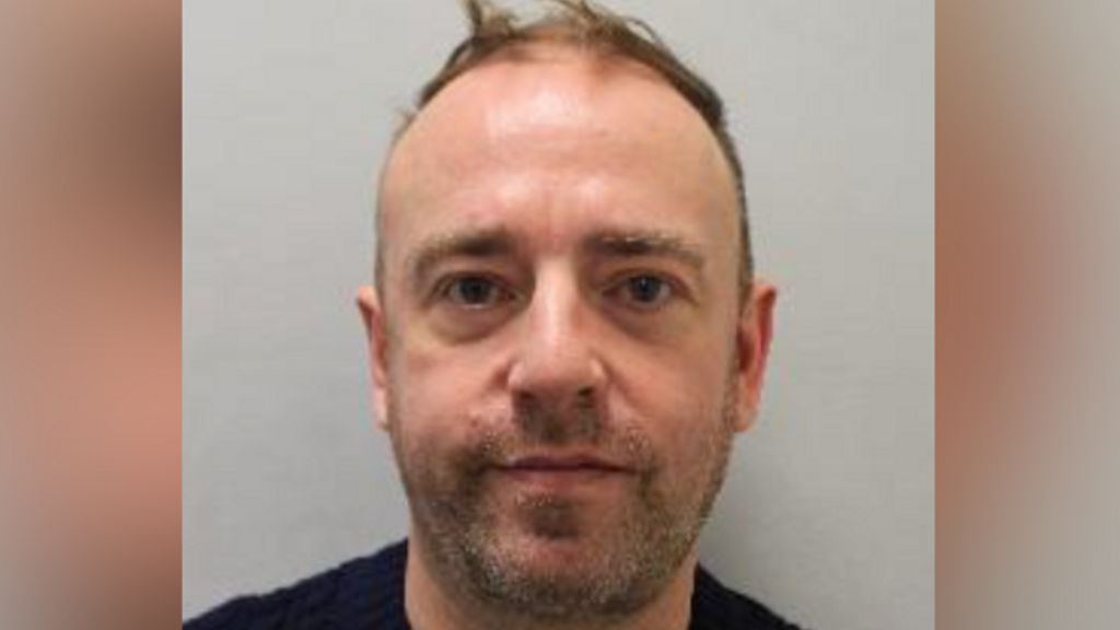 A mug shot of Gunnery who has brown hair and facial stubble and is wearing a black jumper