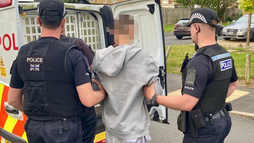 A man wearing a grey tracksuit being escorted by two police officers to the back of a police van