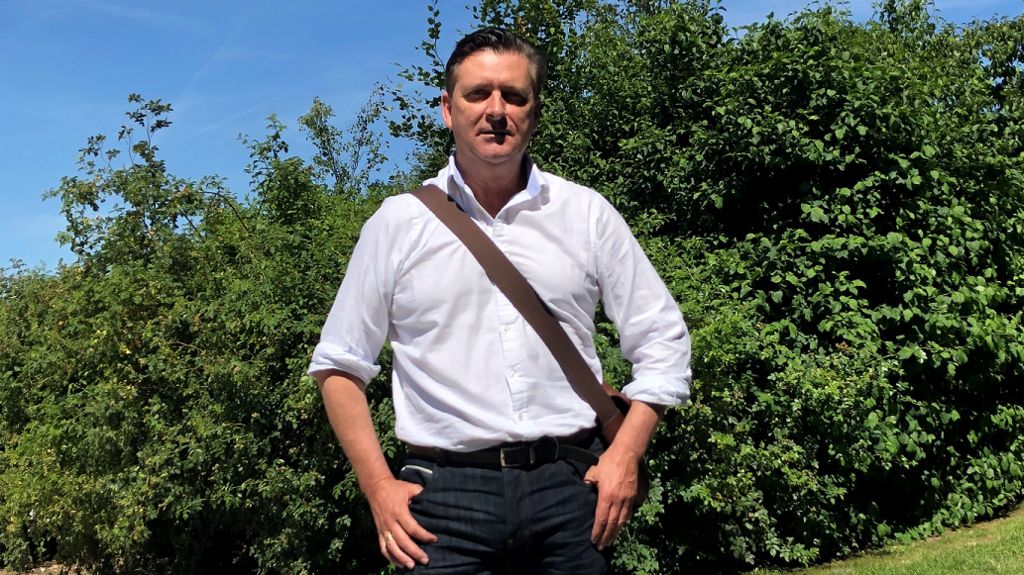 Dr Mark Merrony standing in front of a tree while wearing a white shirt, he has his thumbs looped in his pockets