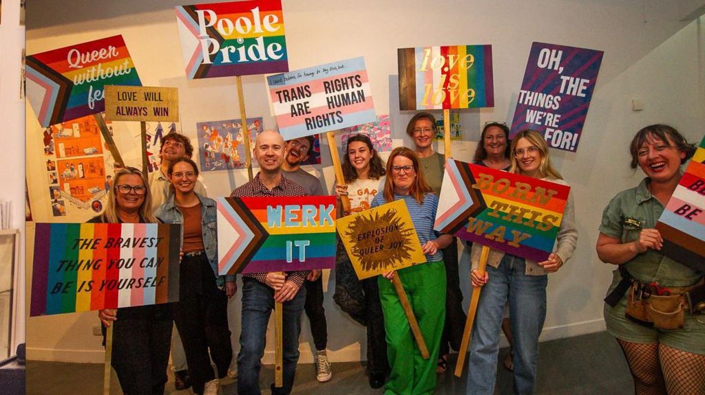 a group of people attending the pride event, mixture of white men and women, holding rainbow coloured protest placards