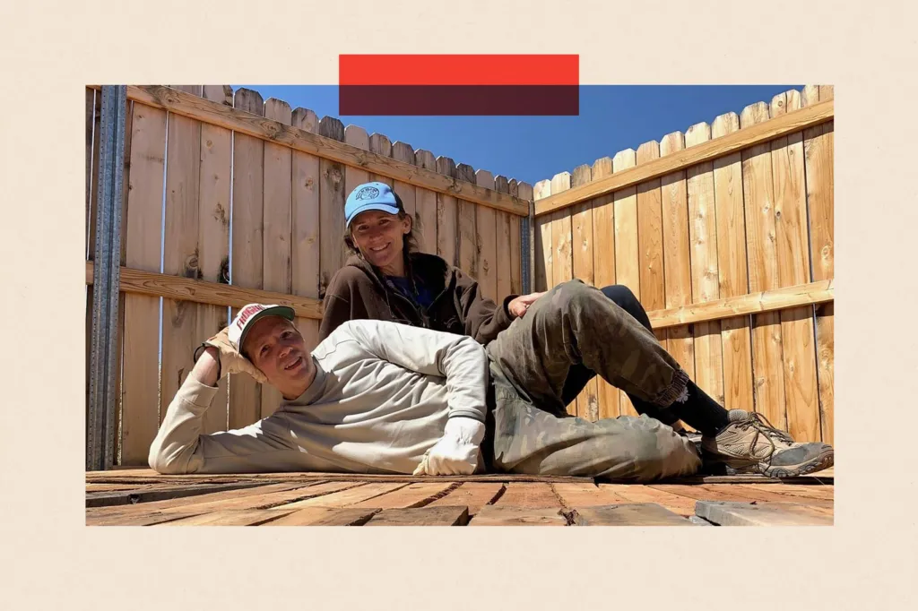 A man and a woman pose on some decking. The man is lying down on the floor, and the woman is sitting behind him. They are both wearing baseball caps and similing.