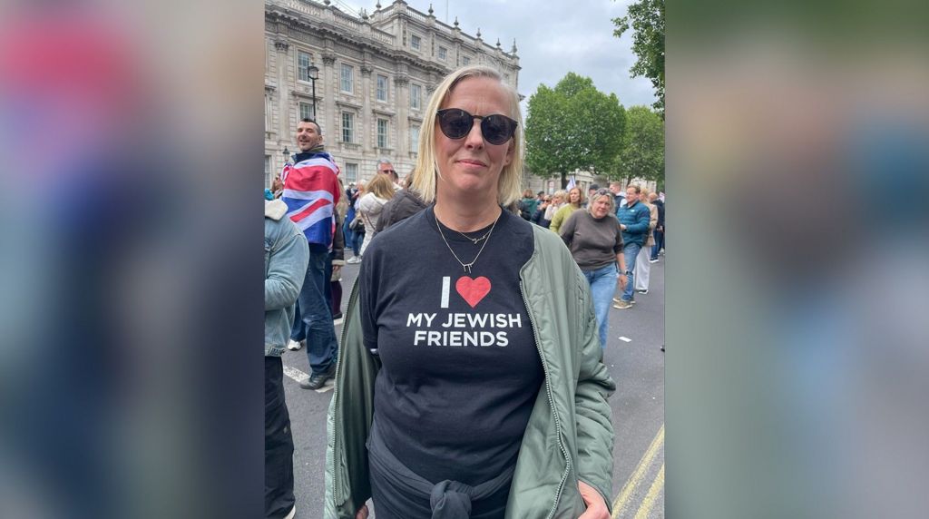 Mary Rendle wears a black tshirt with the words "I love my Jewish Friends", has blonde hair and wears a green coat and black sunglasses. 
She stands in Whitehall with other protestors standing in the background.

